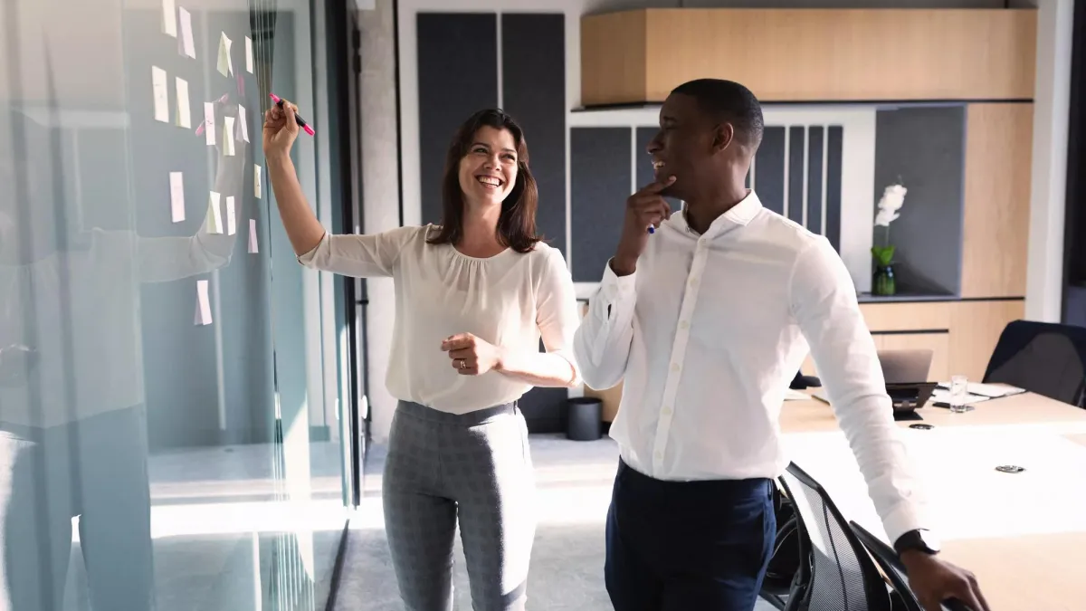 A woman and man brainstorming ideas at a board in an office setting.